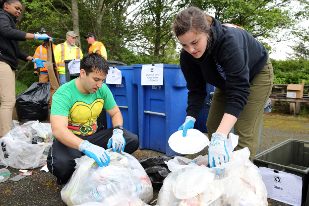 Eine Gruppe von Menschen, die Müll in einem Park sammeln, mit einem Mann und einer Frau in der Mitte, die Handschuhe tragen und Schilder halten, umgeben von Plastikabdeckungen, Flaschen und anderem Müll, einem Mülleimer und einer Holzbank rechts und Bäumen und einem klaren blauen Himmel im Hintergrund.