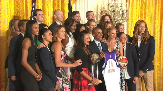 Präsident Obama und First Lady Michelle Obama posieren mit dem Damen-Basketball-Team im Oval Office des Weißen Hauses, halten einen Basketball, Pokal und lächeln in die Kamera neben einer Flagge, Vorhängen und einem Kerzenleuchter.