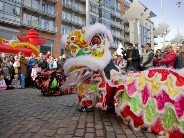 Ein lebhaftes chinesisches Neujahrsfest in Amsterdam mit einem Löwen tanzen im Vordergrund, einer Menschenmenge und Gebäuden im Hintergrund unter einem klaren blauen Himmel.