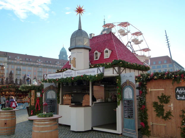 Kleines rotes Dachhaus mit einem Riesenrad davor, umgeben von festlichen Oktoberfest-Dekorationen, Menschen und Gegenständen, vor Gebäuden, Bäumen und einem bewölkten Himmel in München, Deutschland.