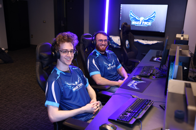 Zwei Männer mit Headsets lächeln, während sie an einem Schreibtisch mit Computermonitoren, Tastaturen und anderer Ausrüstung sitzen und scheinbar ein Videospiel in einem Raum mit einer Wandmontage und Lichtern spielen.