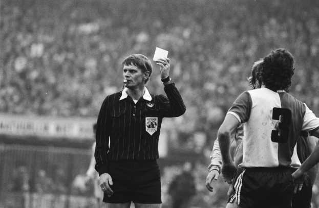 Schwarzes und weißes Bild eines Schiedsrichters mit roter Karte vor einer Menge von Menschen in Fußballtrikots und einem Zaun im Hintergrund.