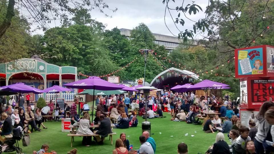 Eine große Gruppe von Menschen sitzt auf einem Rasenfeld mit Schirmen, Tischen und einer Tafel, die ein Biergarten in London anzeigt.