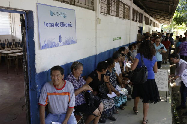 Menschen sitzen auf Bänken vor einem Gebäude, einige halten Papiere und Taschen, mit einer Tafel an der Wand, Tischen und Stühlen links daneben und Bäumen und einem Schuppen im Hintergrund.