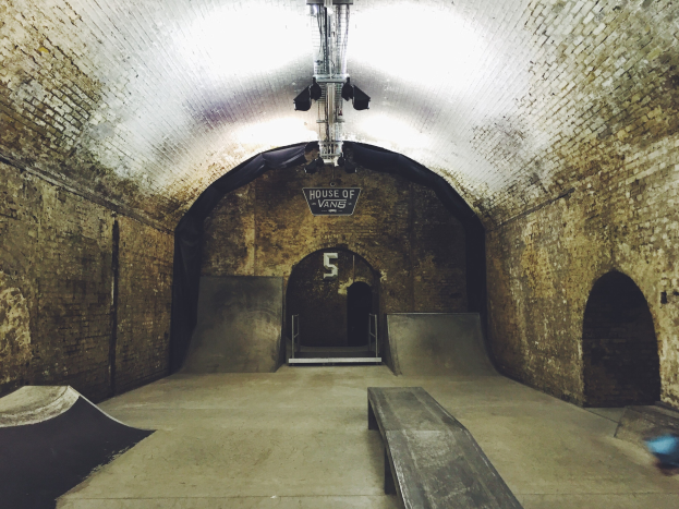 Skatepark mit Backstein-Tunnel, Rampe, Bank und beleuchteten Lampen, mit einer Texttafel oben drauf.