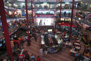 Ein belebtes Einkaufszentrum mit Säulen, Menschen, Tischen, gefliesten Böden, Fahnen, Geschäften, einem Banner, Treppen und einem Namensschild.