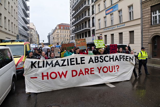 Eine Gruppe von Menschen hält ein Transparent mit der Aufschrift "Klimaziele Abschaffen - Wie wagst du es?" und geht eine Straße entlang mit Fahrzeugen, Gebäuden, Schildern und einem klaren blauen Himmel im Hintergrund.