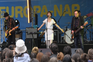 Eine Bühne mit vier Musikern: eine Frau in einem weißen Kleid singt in der Mitte, zwei Männer spielen Gitarren auf der linken und rechten Seite und eine Menge ist unten zu sehen, mit einer Wand und Lautsprechern im Hintergrund.