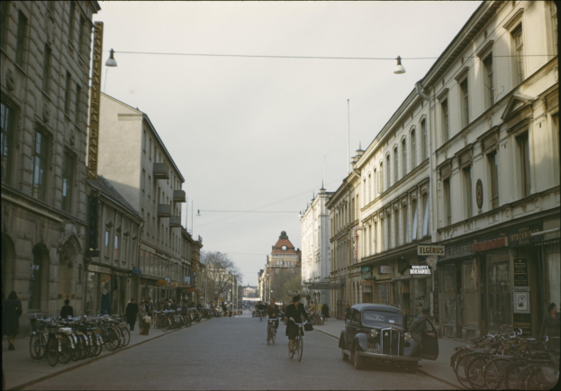 Altes Schwarz-Weiß-Foto einer Stadtstraße mit Fußgängern, Fahrrädern, geparkten Autos, Gebäuden, Bäumen und einem klaren blauen Himmel.
