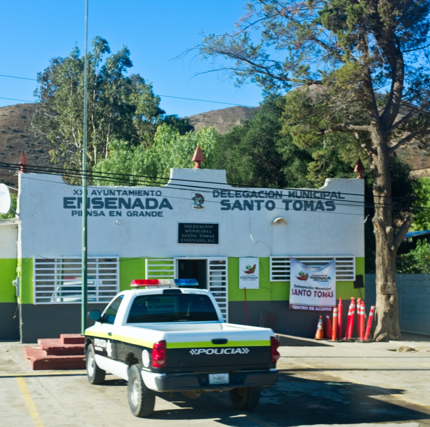 Polizeiauto vor einem Gebäude mit einem Schild, Fenstern, Tür und einer Fahne geparkt, mit Verkehrspfosten, einem Straßenpfosten, einer Schüsselantenne, Kabeln, Bäumen, Hügeln und einem bewölkten Himmel im Hintergrund.