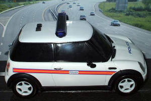 Ein weißer Polizeiwagen steht im Vordergrund mit mehreren fahrenden Fahrzeugen auf der Straße dahinter, umgeben von Gras und Bäumen auf beiden Seiten.