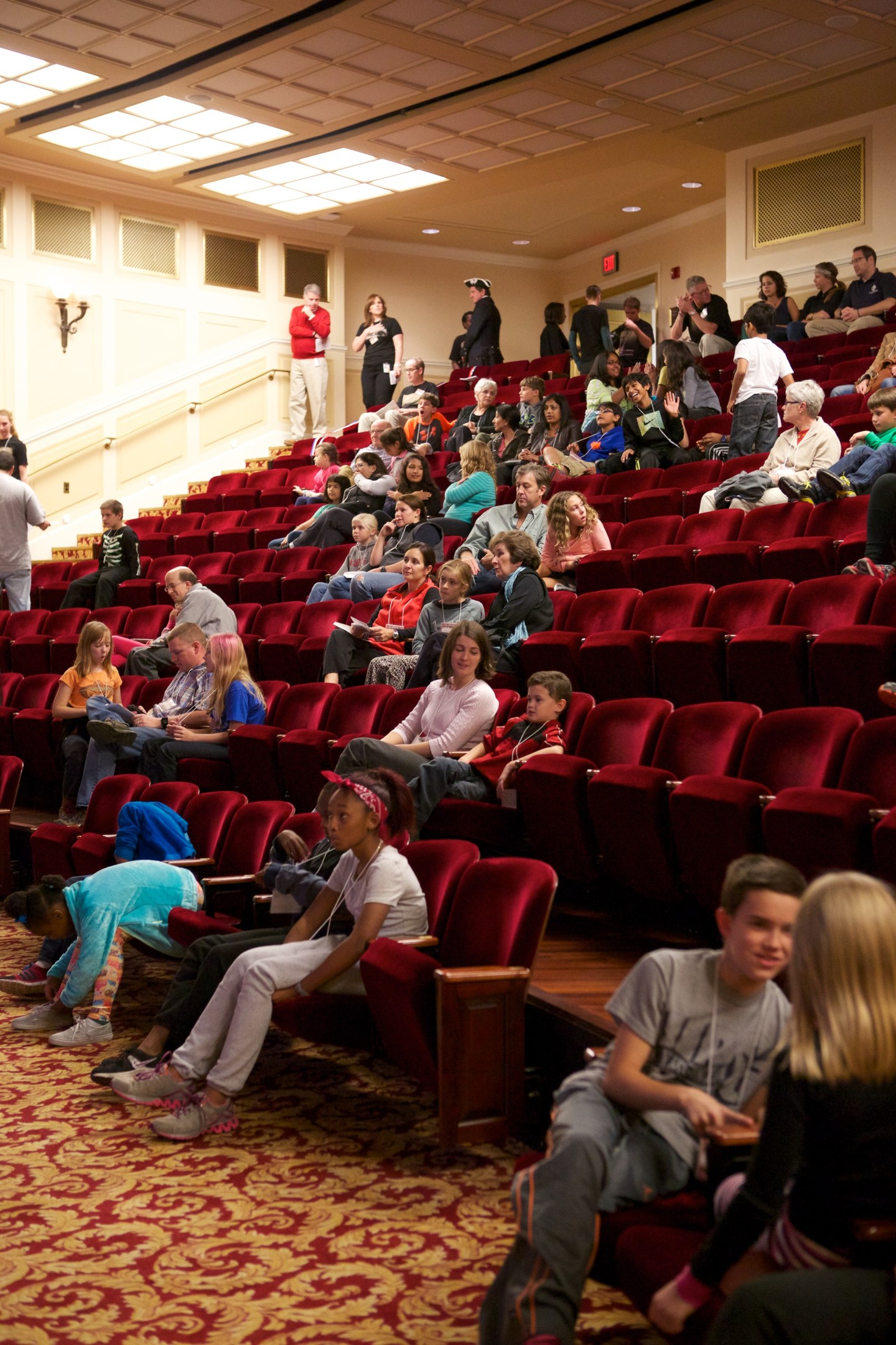 Eine große Gruppe von Menschen sitzt und steht in einem Theater mit verstreuten Stühlen, einer beleuchteten Wand im Hintergrund und Deckenlampen darüber.