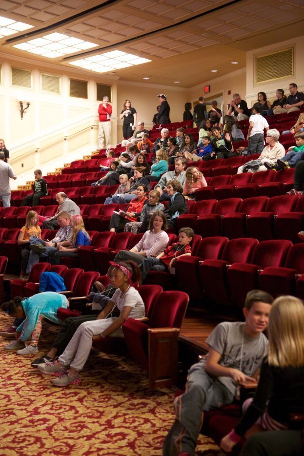 Eine große Gruppe von Menschen sitzt und steht in einem Theater mit verstreuten Stühlen, einer beleuchteten Wand im Hintergrund und Deckenlampen darüber.