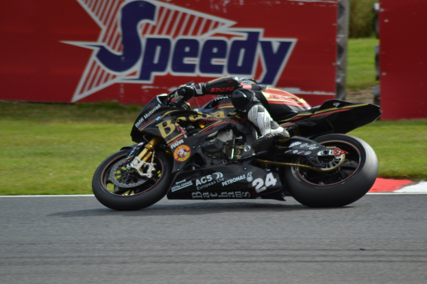 Eine Person mit Helm und Schutzkleidung auf einem Motorrad auf einer Rennstrecke mit Gras und einem Textschild im Hintergrund.