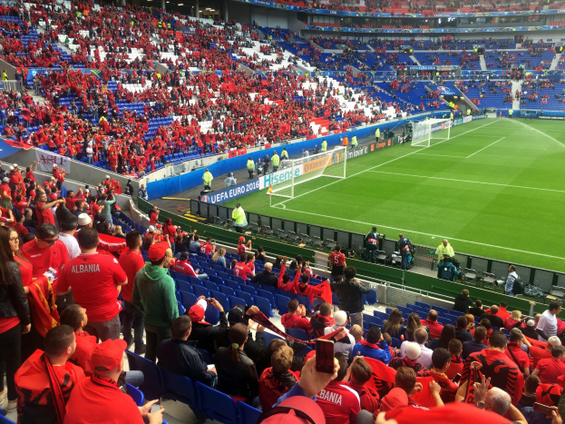 Eine große Menge Menschen in einem hell erleuchteten Stadion schaut ein Fußballspiel zu, mit sitzenden und stehenden Zuschauern, Bannern, einem Torpfosten und einer Decke mit Einbaulichtern.