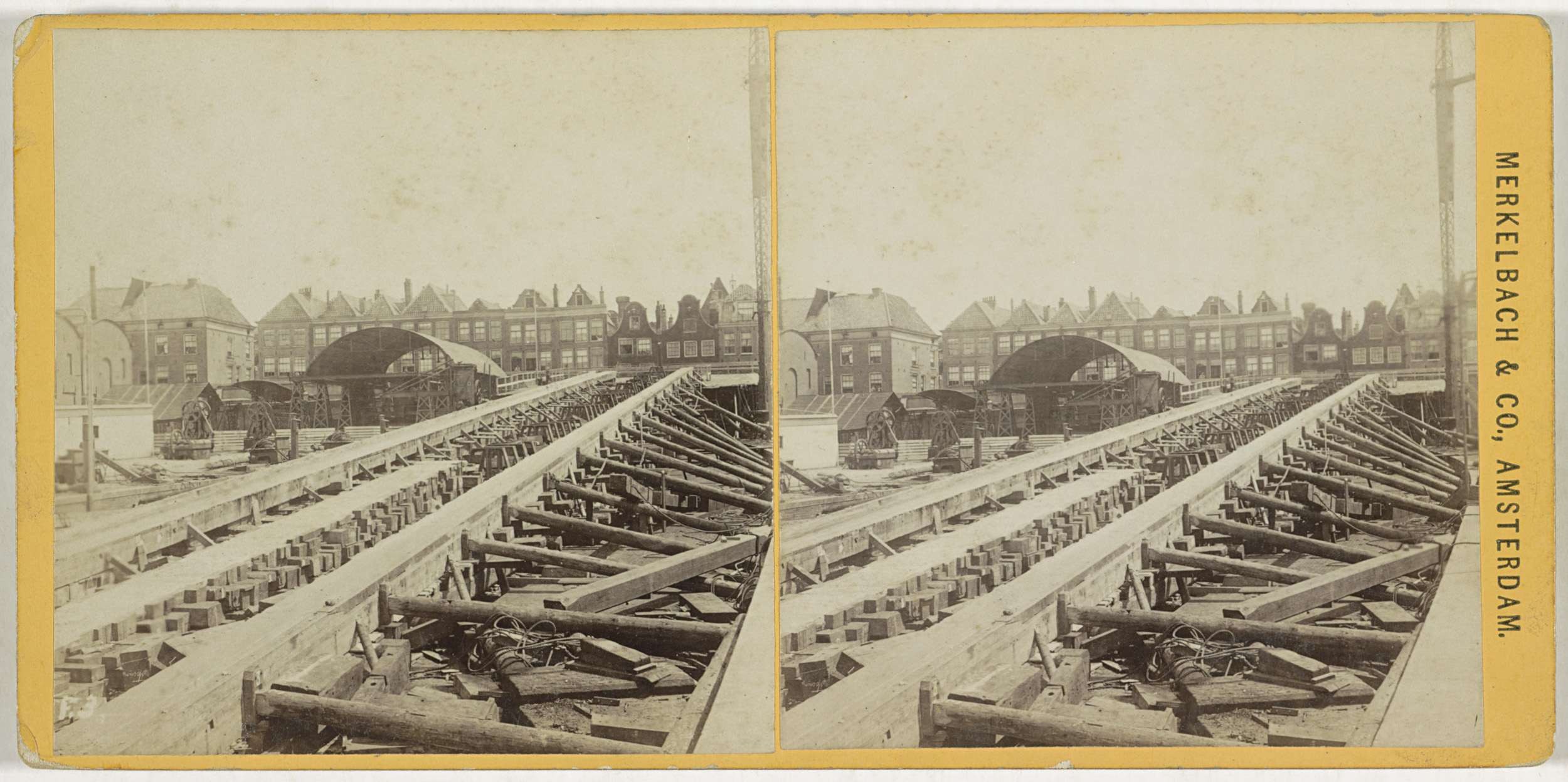 Schwarz-weiß-Foto einer Zugschiene mit Gebäuden im Hintergrund, Text 'Merkelbach & Co, Co. Amsterdam' auf der rechten Seite und verstreute hölzerne Gegenstände im Vordergrund.
