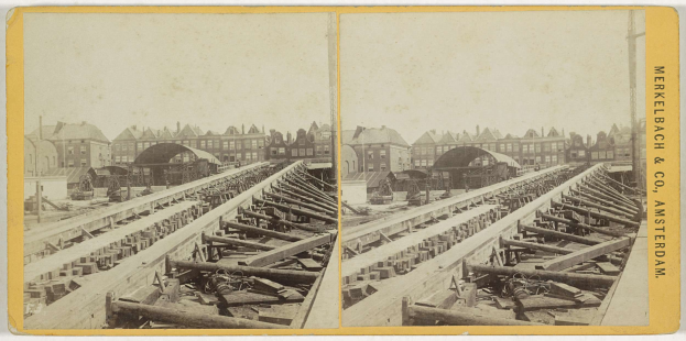 Schwarz-weiß-Foto einer Zugschiene mit Gebäuden im Hintergrund, Text 'Merkelbach & Co, Co. Amsterdam' auf der rechten Seite und verstreute hölzerne Gegenstände im Vordergrund.