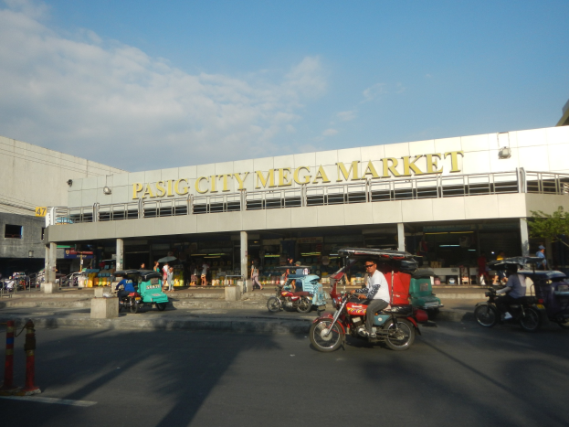 Eine belebte StraßenSzene auf dem Pasig City Mega Market mit Fahrzeugen und Fußgängern, die ein Gebäude mit sichtbarem Text im Hintergrund unter einem bewölkten Himmel zeigen.