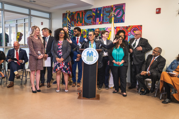 Gruppe von Menschen, die vor einem Rednerpult mit einem Mikrofon und einer Tafel mit der Aufschrift "Sancho-Stadtrat tritt dem Stadtrat bei" stehen, mit sitzenden und stehenden Teilnehmern, Flaggen, einem Feuerlöscher und Deckenleuchten im Hintergrund.