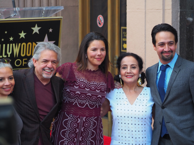 Gruppe von Menschen auf einem roten Teppich bei der Zeremonie des Hollywood Walk of Fame posend, mit einer Kamera auf der linken Seite, einer Tafel mit Text, einem Glaspodest und einer Wand im Hintergrund.