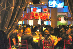 Eine Gruppe von Menschen, die an Tischen mit Essen, Fernsehern, Lampen, hängenden Papieren und Vorhängen in einem Raum sitzen.