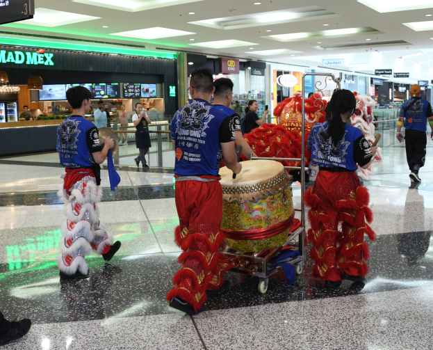 Eine Gruppe von Menschen in roter und blauer Kleidung geht durch einen Flughafen, einige tragen Musikinstrumente und Koffer, mit Geschäften, Namensschildern und Deckenleuchten im Hintergrund, was eine Chinesische Neujahrsfeier in Singapur vermuten lässt.