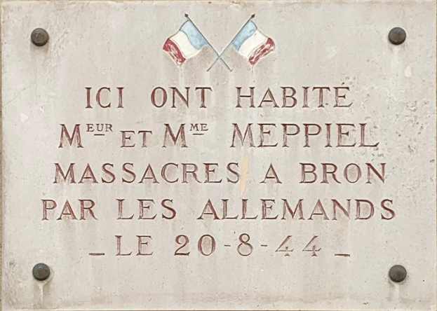 Steintafel an einem Gebäude mit zwei Flaggen darüber, die einen Inschrift tragen, die ein Massaker gedenkt.