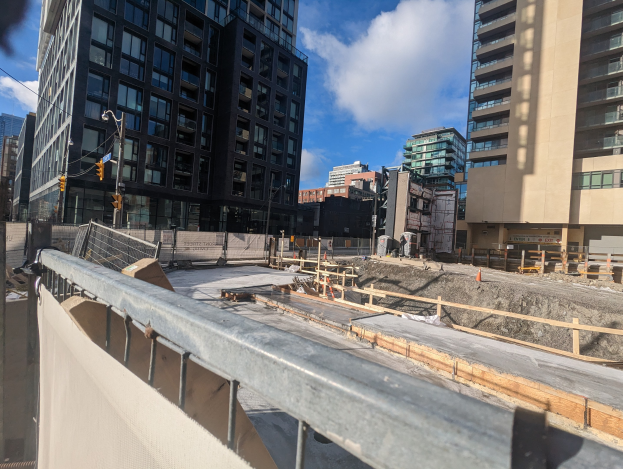 Baustelle in Toronto, Ontario mit einem großen Bauwerk im Bau, umgeben von Absperrungen, Pfählen, Verkehrszeichen und Schildern, unter einem bewölkten Himmel.