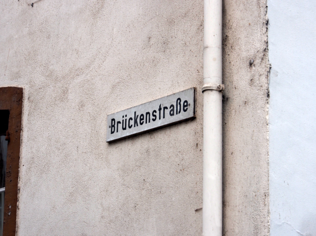 Ein weißes Gebäude mit einem Fenster auf der linken Seite, das ein "Bruckenstraße"-Straßenschild und ein Rohr an seiner Seite zeigt.