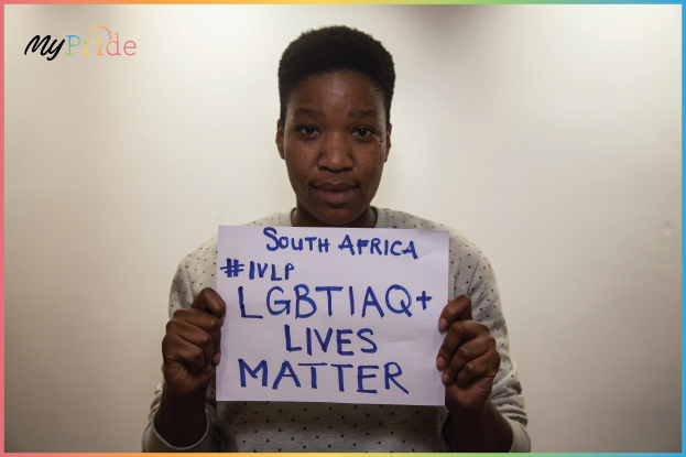 Eine Frau in einem weißen Hemd hält ein Schild hoch, auf dem "LGBTQ+ Lives Matter" steht, mit entschlossener Miene vor einer Wand.
