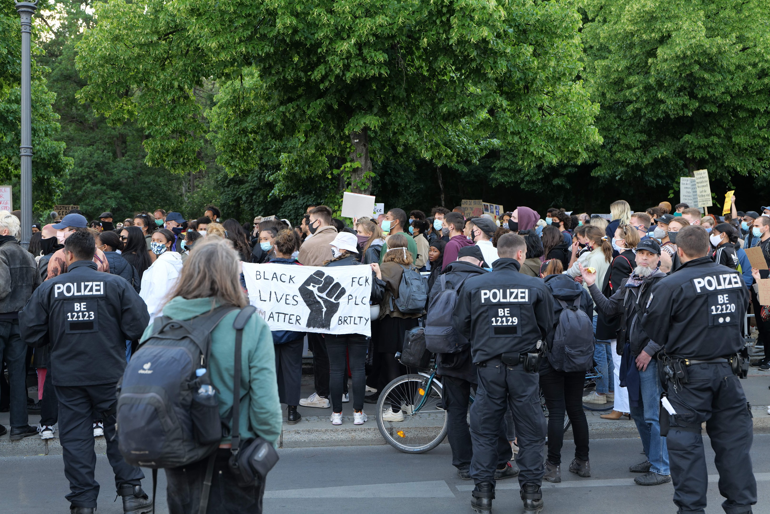 Eine große Gruppe von Menschen bei einer Black Lives Matter Demonstration in Berlin, einige halten Schilder und tragen Kappen und Taschen, mit einem Fahrrad im Vordergrund und Bäumen und einem Pfahl im Hintergrund.