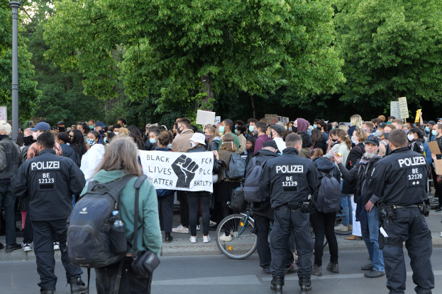 Eine große Gruppe von Menschen bei einer Black Lives Matter Demonstration in Berlin, einige halten Schilder und tragen Kappen und Taschen, mit einem Fahrrad im Vordergrund und Bäumen und einem Pfahl im Hintergrund.