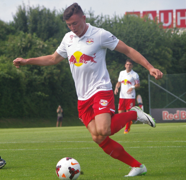 Ein professioneller Fußballspieler in einem roten Trikot kickt einen Ball auf einem Rasenfeld mit Bäumen und einem klaren blauen Himmel im Hintergrund, in der Nähe einer Tafel mit der Aufschrift "RB Leipzig."