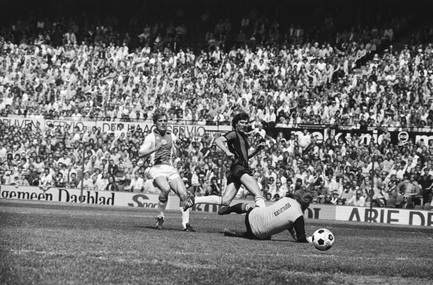 Schwarzes Foto von Männern, die auf einem Feld Fußball spielen, mit Zuschauern im Hintergrund und einer "Bundesliga 1978-79 Ajax 1-1 Hannover 96"-Tafel.