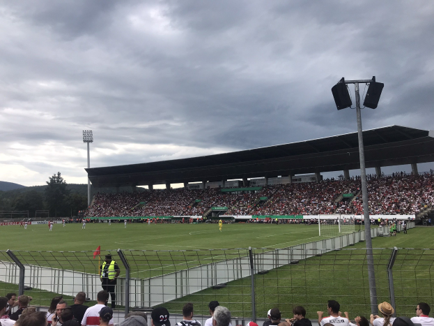 Große Zuschauermenge bei einem Fußballspiel in einem bewölkten, offenen Stadion mit beleuchtetem Feld und umgebenden Bäumen und Hügeln.