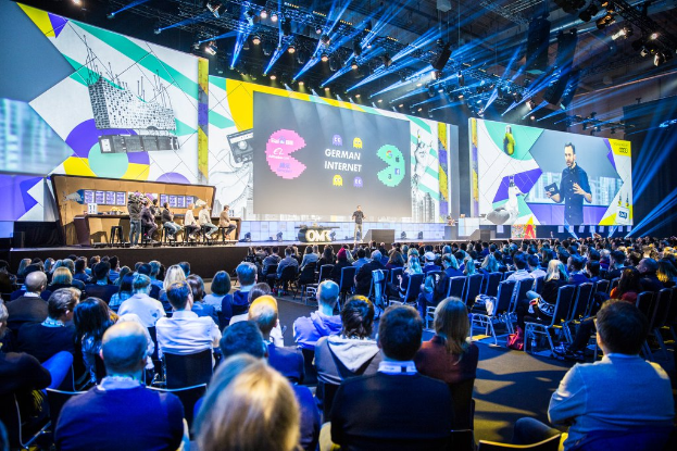 Eine große Menschenmenge, die auf Stühlen vor einer Bühne sitzt, mit einigen Personen, die auf dem Podium stehen, einem Tisch mit verschiedenen Gegenständen links und Bildschirmen und Lichtern im Hintergrund auf der Deutschen Internetkonferenz 2019.