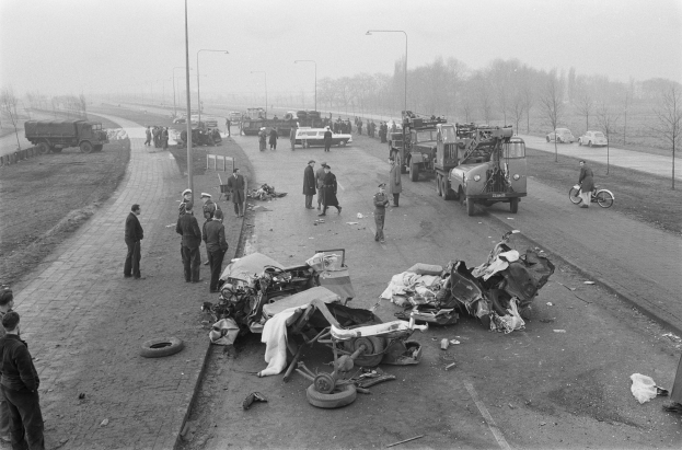 Schwarz-weißes Bild einer Autounfall-Szene am Straßenrand mit mehreren Fahrzeugen, einer Gruppe von Menschen in der Nähe und Hintergrund-Elementen wie Laternenpfähle, Bäume und den Himmel.