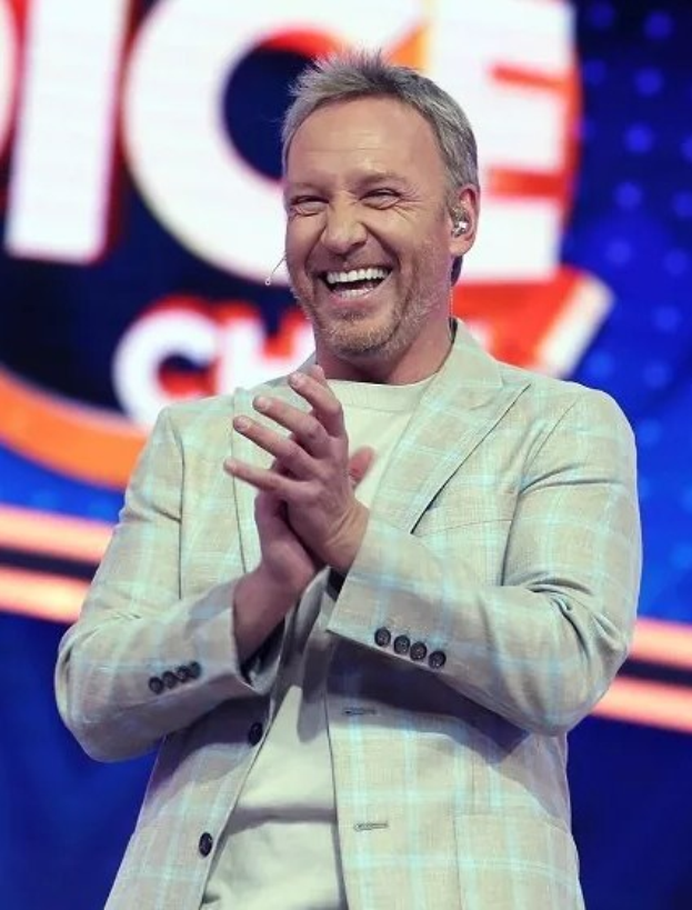 Ein Mann in einem Anzug, der auf einer Bühne klatscht, mit 'The Voice'-Schriftzug im Hintergrund.