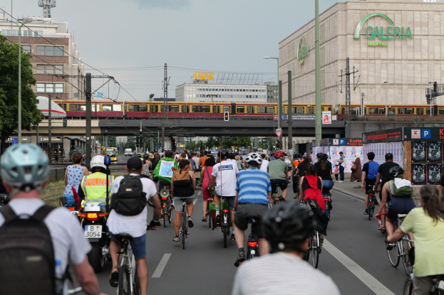 Eine Gruppe von Menschen, die auf Fahrrädern eine Straße entlangfahren, die von hohen Gebäuden gesäumt ist, einige tragen Helme und Taschen, mit einem Zug auf einem Bahngeleis, Strommasten, Bäumen und einem klaren blauen Himmel im Hintergrund.