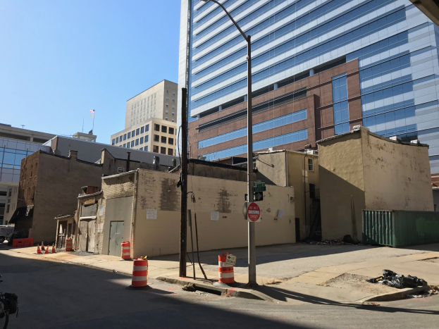 Eine Stadtstraße mit einem geparkten Fahrrad, umgeben von Gebäuden, Straßenmöbeln, Fahrzeugen und einem abgerissenen Gebäude im Hintergrund unter einem klaren blauen Himmel.
