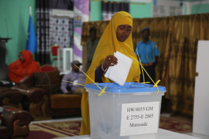 Eine Frau mit gelbem Kopftuch steht an einer Wahllokal, wo sie ihre Stimme vor einem Tisch mit einer Box abgibt, während einige Menschen auf der linken Seite sitzen, mit Vorhängen, einer Flagge und einer Wand im Hintergrund.