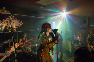 Eine Frau in einer schwarzen Jacke singt in ein Mikrofon auf der Bühne, mit einem Mann in Sonnenbrille und einem Stirnband, der ein Instrument spielt, hinter ihr, unter einem Schild, auf dem "LA Guns" steht und Diskolichter leuchten.
