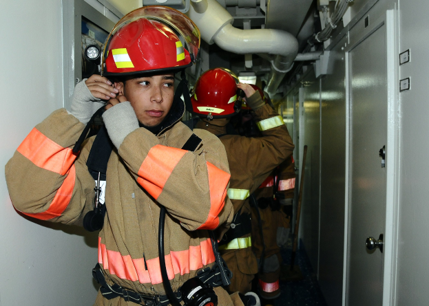 Feuerwehrleute in Uniform stehen in einem Raum mit Rohren und Gegenständen im Hintergrund, wahrscheinlich während einer Übung.