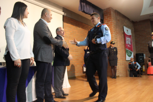 Eine Gruppe von Menschen auf einer Bühne mit einer Person in Uniform, die eine Mütze hält, eine Frau in einem weißen Hemd, ein Banner, ein Lautsprecher auf einem Ständer, Vorhänge, ein Tisch und ein Bildschirm an der Wand im Hintergrund.