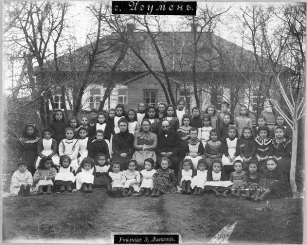 Schwarzes und weißes Foto einer Gruppe von Kindern, die vor einem Gebäude mit Fenstern posen, umgeben von Bäumen und einem klaren Himmel, mit Text unten, der "Jugoslawien, die erste Schule der Welt" liest.