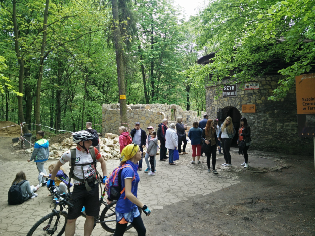 Gruppe von Menschen, die einen bewaldeten Pfad entlanggehen, einige tragen Helme und tragen Taschen oder Fahrräder, mit einer Steinmauer und einem Schild im Hintergrund, das den Eingang zum Szyb Stylweste Mountainbike Trail anzeigt.