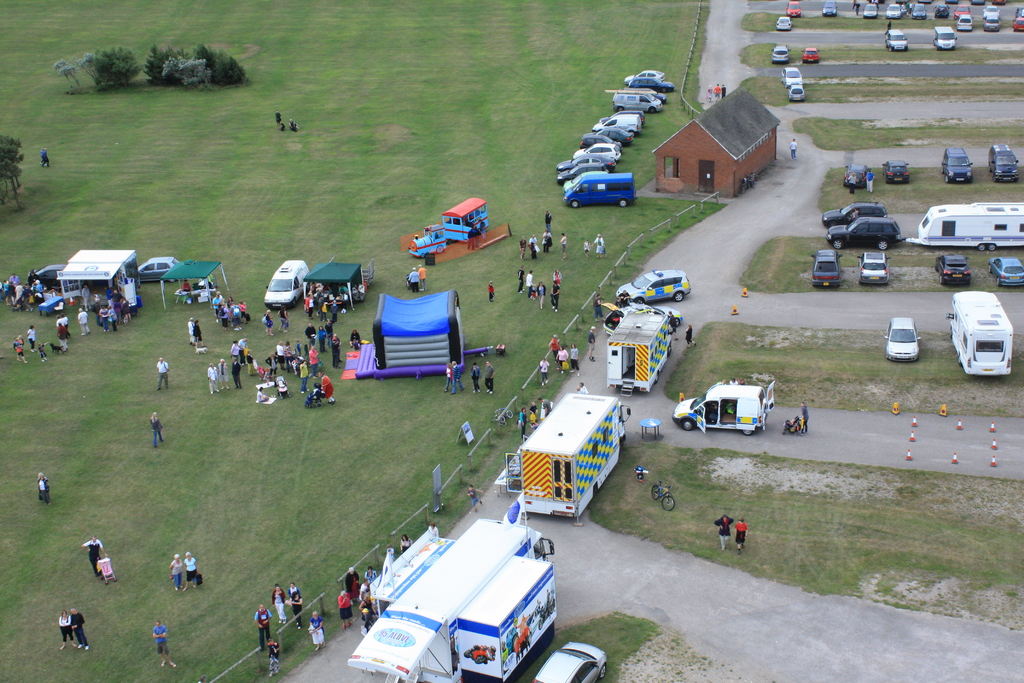Eine Übersicht eines Parkplatzes mit vielen Fahrzeugen, einigen Menschen in der Nähe von Zelten, einer zentralen Hütte und einigen Bäumen auf der linken Seite.