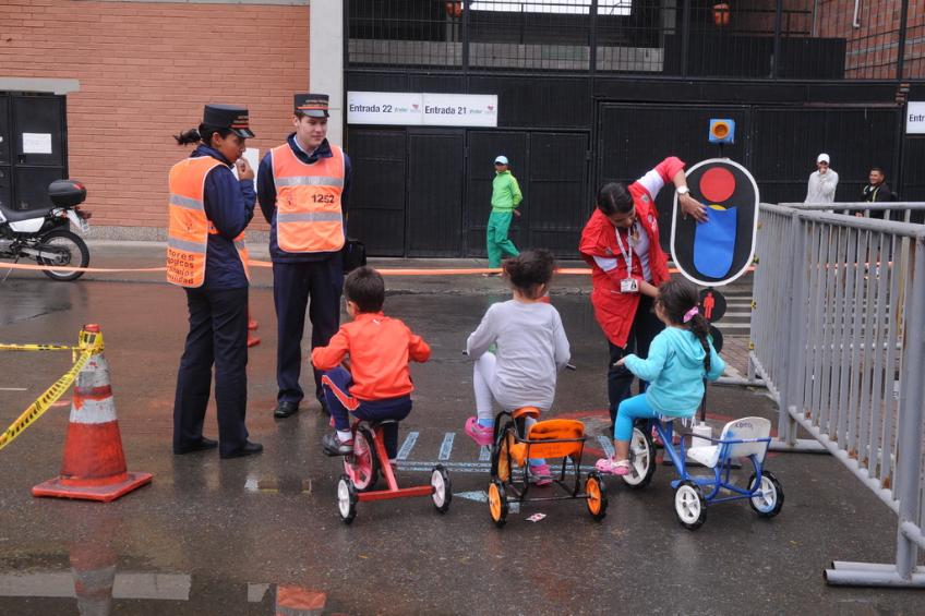 Kinder auf Fahrrädern auf einer Straße mit Verkehrshütchen, Menschen, Geländern, einer Wand mit Türen und Schildern im Hintergrund.