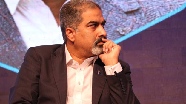 Ein Mann in Anzug sitzt auf einer Bühne und denkt nach vor einem Bildschirm.