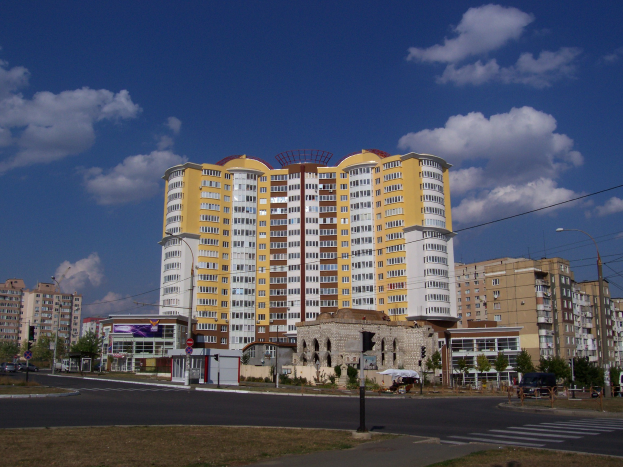 Großes Wohnhaus in einer städtischen Umgebung mit Straßeninfrastruktur, Fahrzeugen, Bäumen und Wolkenhimmel.
